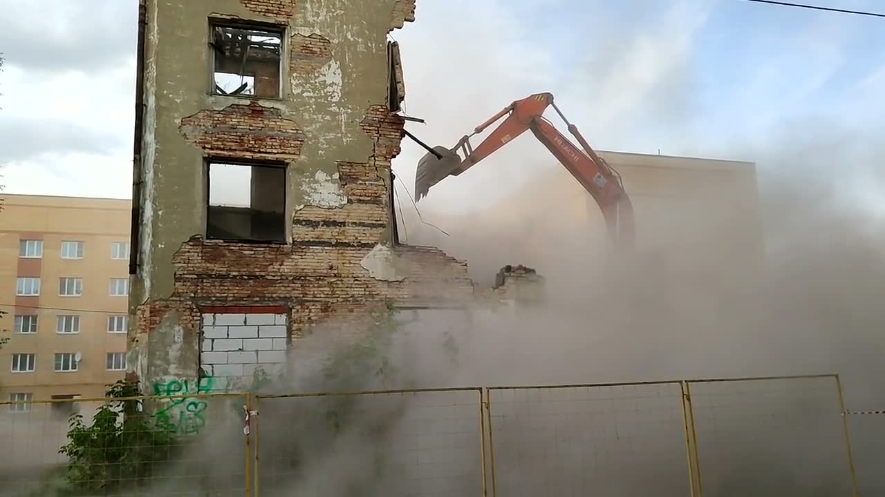 An orange excavator arm tearing into the brick wall of a derelict building, dust billowing around it